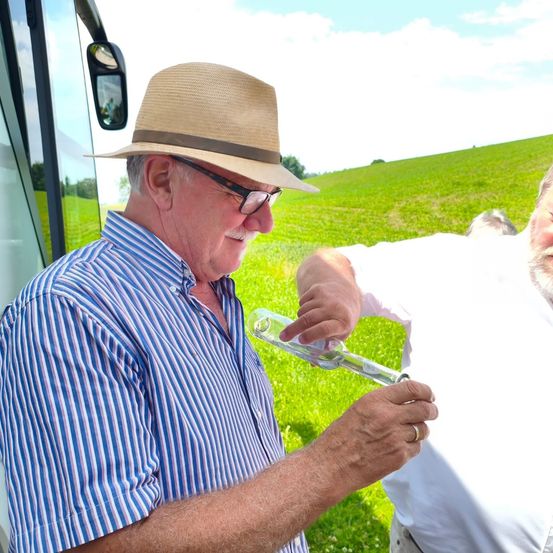 Ein älterer Mann in einem blau gestreiften Hemd und einem Strohhut trinkt aus einer Flasche und steht in einem grünen Feld. Ein anderer Mann hilft ihm. Hinter ihnen ist ein Fahrzeug geparkt.
