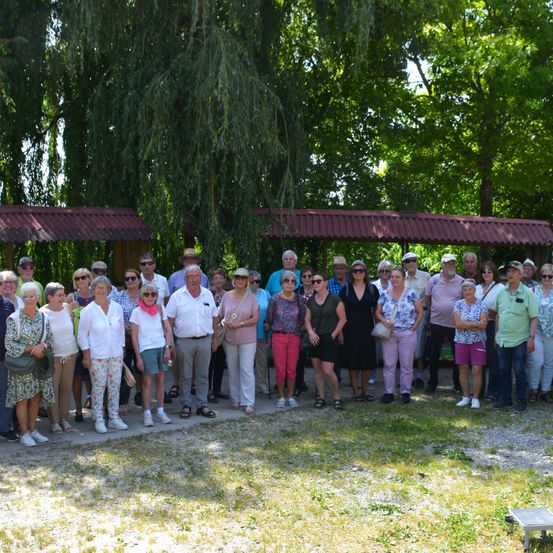 Eine Gruppe von Menschen in Sonnenbrillen und Hüten steht draußen, mit Bäumen und roten Dächern im Hintergrund. Einige tragen Brillen, Schals und Armbänder. Sie sind wahrscheinlich auf einer Klassenfahrt.