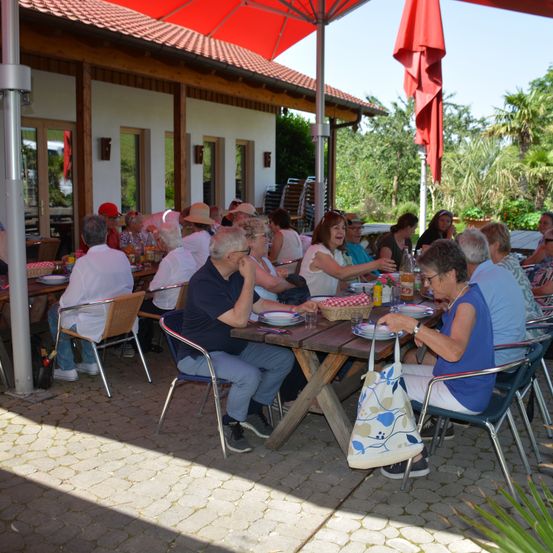 Eine Gruppe von Menschen isst draußen in einem Restaurant. Sie sitzen an Tischen unter roten Sonnenschirmen. Die Tische sind mit Tellern, Servietten und verschiedenen Getränken gedeckt. Im Hintergrund sind Bäume und ein Gebäude zu sehen.