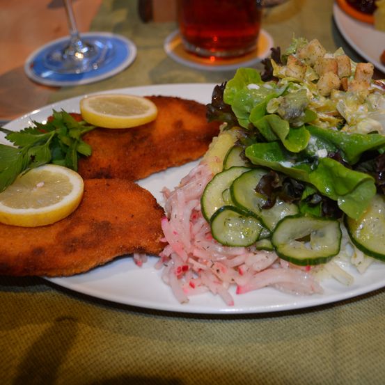 Ein Teller mit gebratenem Fleisch, Zitronenscheiben, Petersilie und einem Salat mit Gurken und Croutons. In der Nähe steht ein Glas dunkles Bier.