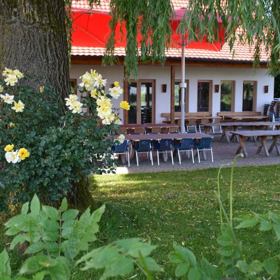Ein Restaurantgarten mit draußen aufgestellten Tischen und Stühlen. Der Garten hat gelbe Blumen und einen roten Regenschirm. Das Gebäude hat Glastüren und Fenster.