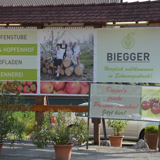 Ein Schild zeigt Bilder von Obst und Menschen. Es steht 'Biegger' und 'Herzlich willkommen in Schwarzenbach!' Eine Familie posiert neben einem Holzstapel. Frische Kirschen werden unten beworben.