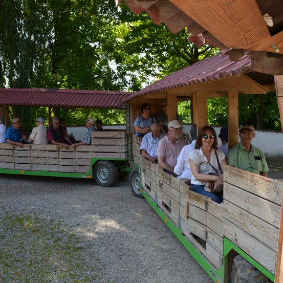 Mehrere Menschen sitzen auf einem hölzernen, von einem Traktor gezogenen Wagen an einem sonnigen Tag. Einige tragen Hüte und andere Sonnenbrillen. Sie scheinen auf einer Tour zu sein.