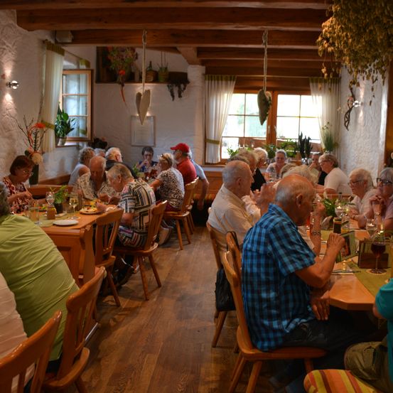 Eine Gruppe älterer Menschen sitzt an Tischen in einem Restaurant. Der Raum hat Holzböden und -wände, mit hängenden Pflanzen und einer herzförmigen Dekoration. Einige essen, während andere sprechen.