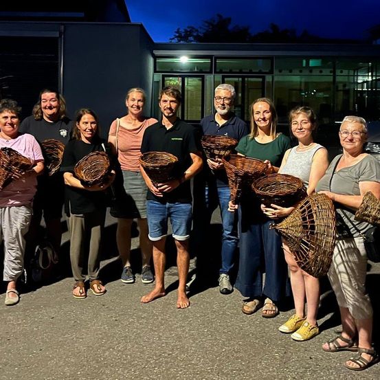 Eine Gruppe von Menschen, die geflochtene Körbe halten, posiert für ein Foto draußen bei Nacht. Sie stehen in einer Reihe, mit einem Gebäude im Hintergrund.