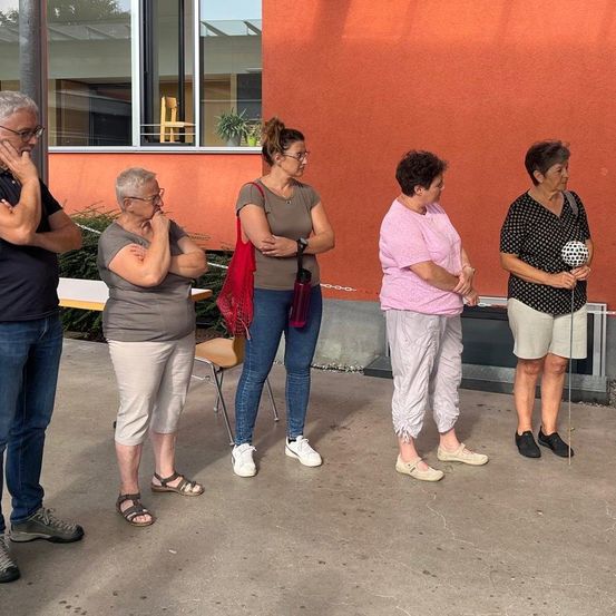 Eine Gruppe älterer Erwachsener, alle Frauen, stehen in einer Reihe vor einem orangefarbenen Gebäude. Sie tragen Freizeitkleidung und Sneakers.