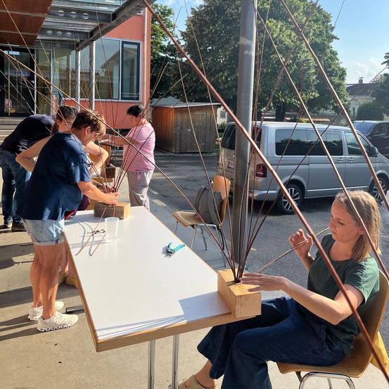 Eine Gruppe von Menschen arbeitet draußen an einem Projekt, eine Frau sitzt auf einem Stuhl, während eine andere Frau ihr hilft. Sie sind umgeben von Holzstäben, einem Tisch und Stühlen. Ein Van ist in der Nähe geparkt.