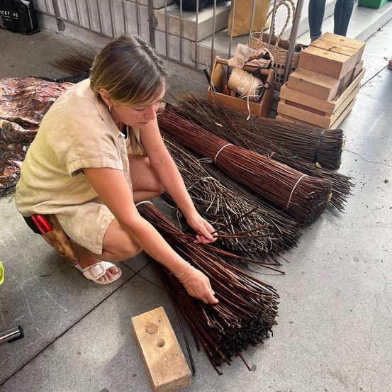 Eine Frau arbeitet auf dem Boden sitzend an einem Bündel brauner Zweige. Ein Holzblock steht neben ihr. Hinter ihr befinden sich mehrere Holzkisten und Körbe. Der Boden ist aus Beton.