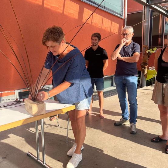 Eine Frau arrangiert eine Pflanze in einem braunen Block auf einem Tisch. Ein Mann mit Brille und ein weiterer Mann beobachten sie. Eine Frau steht neben ihnen. Die Wand hinter ihnen ist orange gestrichen.