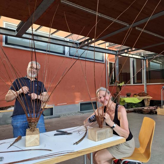 Ein erwachsener Mann und eine Frau fertigen Objekte aus dünnen Zweigen an, die wie Körbe aussehen, sitzend an einem Tisch mit Werkzeugen. Hinter ihnen steht ein rotes Gebäude mit Glasfenstern.