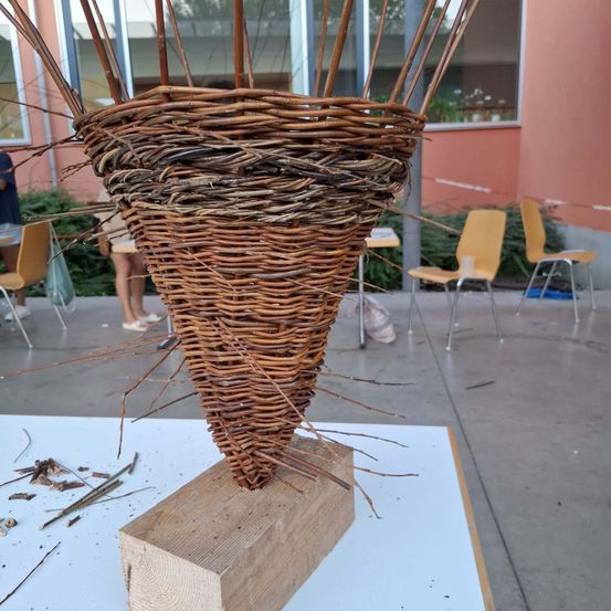 Ein Weidenkorb aus Zweigen ist auf einem Holzblock in einer Außenumgebung platziert. Im Hintergrund sind Menschen in der Nähe eines Gebäudes mit Glasfenstern zu sehen.