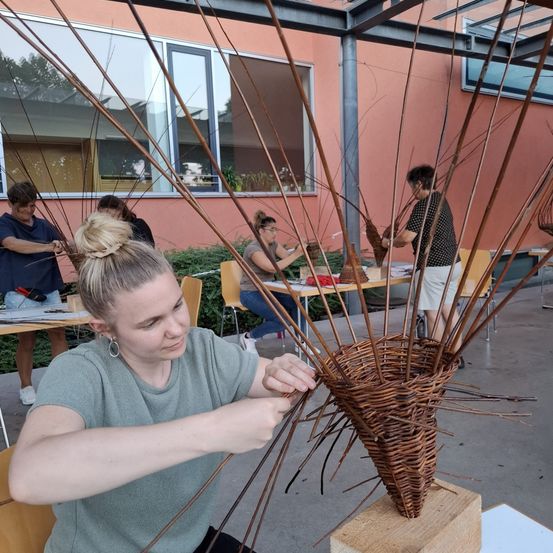 Eine Frau fertigt einen Korb mit konischer Form aus Weidenzweigen an, während mehrere Personen ähnliche Projekte in einer Werkstatt bearbeiten.