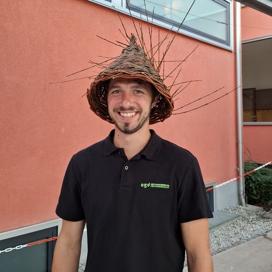 Ein Mann in einem schwarzen Poloshirt mit Logo und einem geflochtenen Hut mit Zweigen steht vor einem rosa Gebäude.