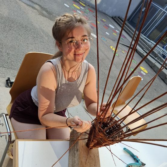 Eine junge Frau mit Brille sitzt und webt draußen einen Korb. Sie sitzt auf einem Stuhl und hält ein Stück Holz.