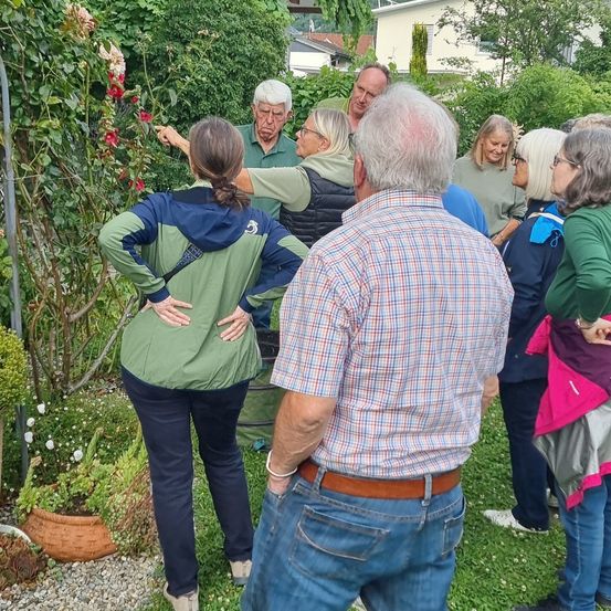 Eine Gruppe von Menschen, darunter ein älterer Mann, versammelt sich in einem Garten und untersucht Pflanzen und Blumen. Sie sind alle lässig gekleidet.