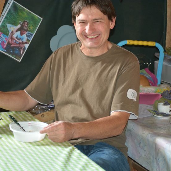 Ein lächelnder Mann sitzt an einem Tisch mit einer Schüssel, umgeben von einer blumigen Tischdecke und einer Tasse mit Lavendel.