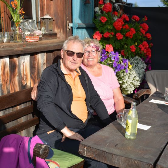 Ein älteres Paar mit Brille und lächelnd, das auf einer Bank vor einer hölzernen Bar sitzt, mit bunten Blumen im Hintergrund.