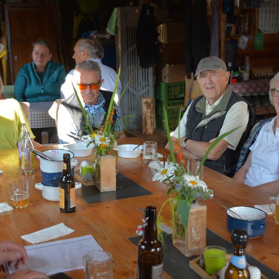 Vier Personen sitzen um einen Tisch mit Blumen und Bierflaschen. Eine Person trägt eine Brille.