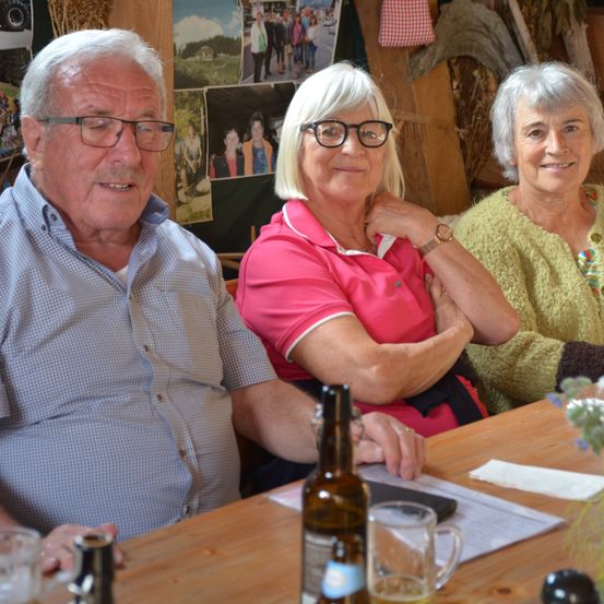 Drei ältere Personen sitzen lächelnd an einem Holztisch. Der Mann trägt eine Brille und ein blaues Hemd. Die Frauen tragen rosa und gelbe Pullover. Hinter ihnen zeigt eine Wand verschiedene Fotos.