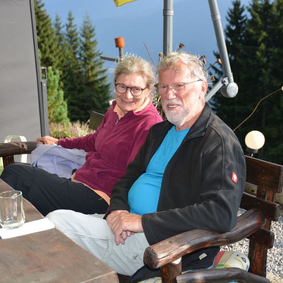 Ein älteres Paar sitzt lächelnd auf einer Bank. Dahinter ist eine Landschaft mit Bergen und Bäumen. Ein Glas steht auf dem Tisch vor ihnen.