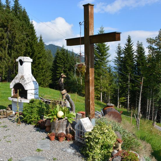 Ein Holzkreuz steht auf einem Kiesweg mit Pflanzen, einem weißen Ofen und einem Plüschtier. Im Hintergrund befindet sich ein grasbewachsener Hügel mit Bäumen und einem bewölkten Himmel.