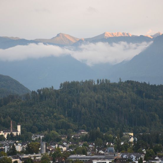 Eine bergige Landschaft mit einer Stadt darunter, eingehüllt in Morgennebel, zeigt einen großen Hügel mit dichten Bäumen und einem goldenen Sonnenuntergang am Horizont.