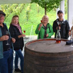 Eine Gruppe von fünf Personen, die lächeln und Weingläser halten, steht um ein großes Holzfass. Sie sind draußen mit einem Weinberg im Hintergrund.
