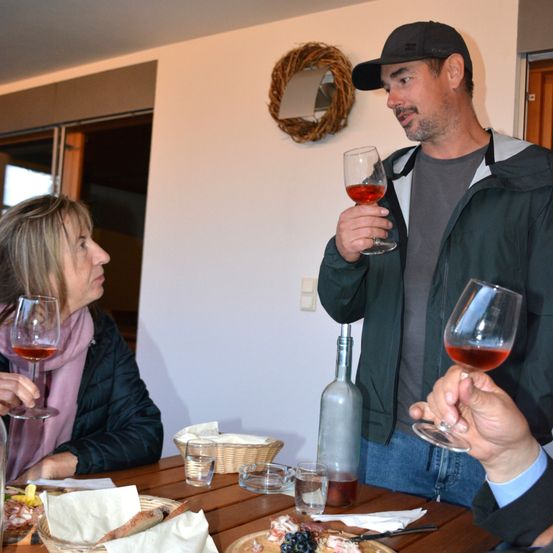 Eine Gruppe von Menschen sitzt an einem Tisch, mit Weingläsern in den Händen. Ein Mann steht und hält ein Weinglas mit rotem Wein. Es gibt einen Korb mit Brot und anderen Speisen auf dem Tisch.