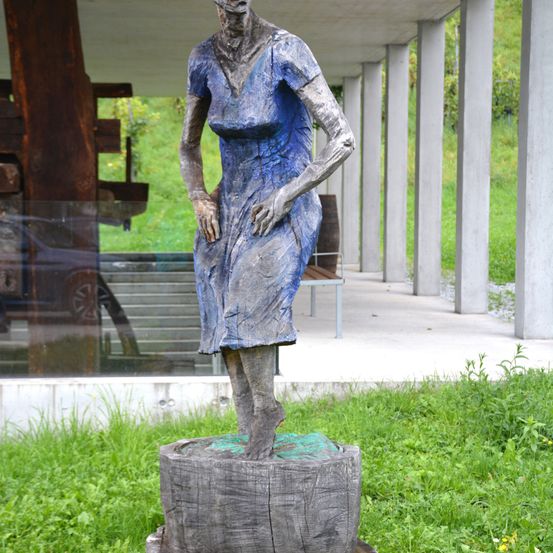 Eine blaue Statue einer Frau steht auf einem Holzstumpf im Freien. Sie trägt ein Kleid und hat die Hände auf die Hüften. Dahinter steht eine moderne Struktur mit Säulen und einer Holzbank.