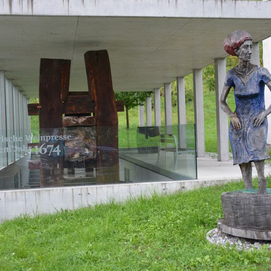 Eine Statue einer Frau steht auf einem Rasen vor einem modernen Gebäude. Die Frau trägt ein blaues Kleid und eine rote Kopfbedeckung. Dahinter befindet sich eine hölzerne Struktur und eine Glaswand mit Reflexionen.
