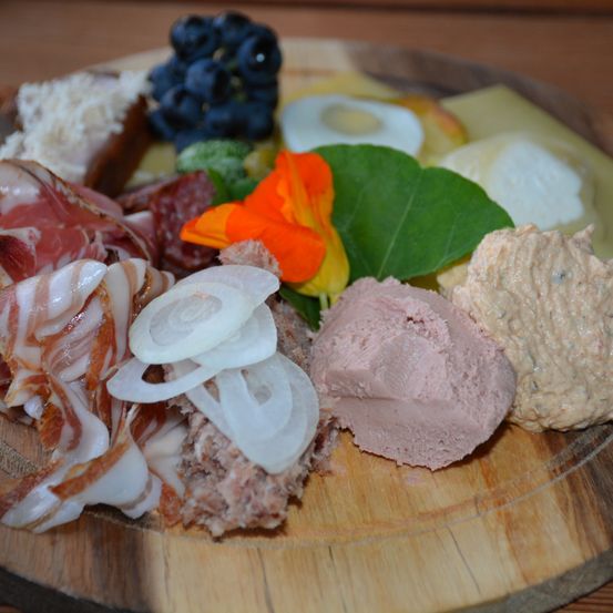 Ein Holzteller präsentiert eine Auswahl an Lebensmitteln, darunter geschnittene Fleischsorten, Käse, Trauben, Eier, Zwiebeln und Blumen.