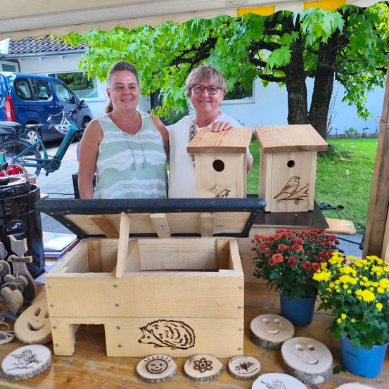 Zwei Frauen stehen hinter einem Holztisch mit Vogelhäusern, Kunsthandwerk und Blumen in einer sonnigen Außenumgebung.