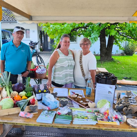 Drei Personen stehen hinter einem Tisch voller Gemüse und Werbematerialien in einer Außenumgebung. Sie lächeln und scheinen an einem Marktereignis teilzunehmen. Hinter ihnen steht ein Haus mit einem geparkten Fahrrad.
