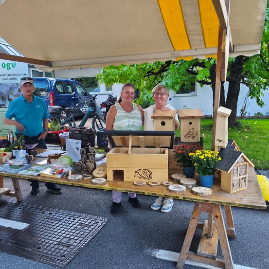 Drei Personen stehen hinter einem mit einem Zelt bedeckten Tisch. Sie lächeln und posieren für ein Foto. Auf dem Tisch befinden sich hölzerne Vogelhäuschen, Blumen und andere Gegenstände. Dahinter steht ein Baum und ein Gebäude.