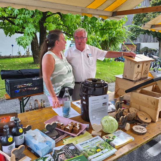 Ein Mann und eine Frau stehen an einem Tisch im Freien und präsentieren Gartenprodukte. Der Tisch ist mit Büchern, Holzkisten und einem Kohl geschmückt. Sie stehen unter einem Zelt, mit einem Baum und Fahrrädern im Hintergrund.