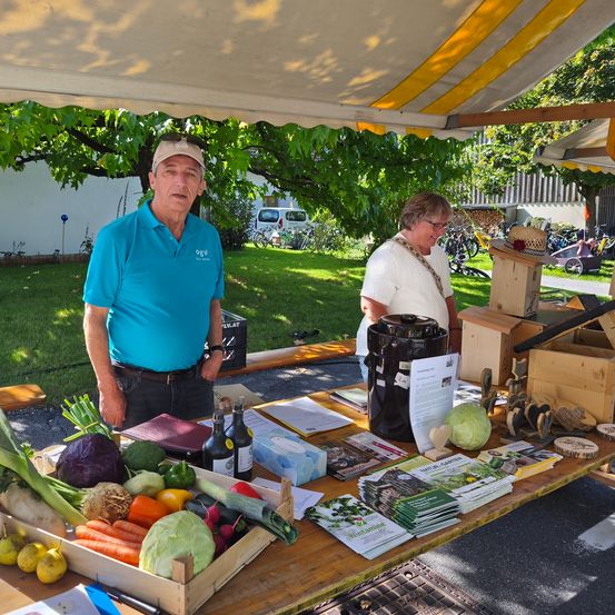 Ein Mann und eine Frau stehen unter einem Vordach neben einem Tisch mit Gemüse, Flaschen und Broschüren. Der Mann trägt ein blaues Poloshirt, während die Frau ein weißes Shirt und eine Brille trägt. Hinter ihnen sind Fahrräder in der Nähe eines Baumes geparkt.
