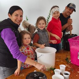 Eine Familie in einem Workshop stellt etwas her. Eine Frau hält einen Eimer, und Kinder und ein Mann schauen zu. Tassen und ein rosa Behälter stehen auf dem Tisch.