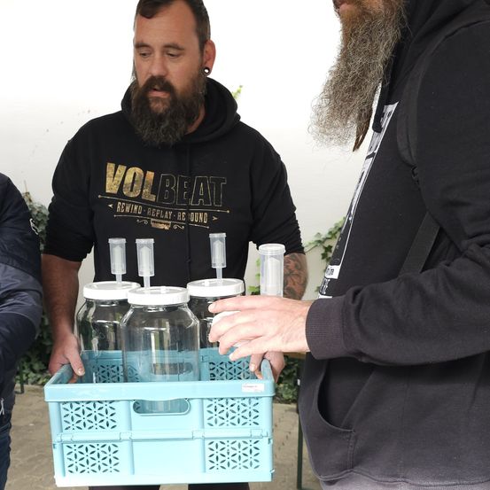 Zwei bärtige Männer in schwarzen Sweatshirts halten einen blauen Korb mit mehreren klaren Gläsern, jedes mit einem weißen Deckel und einem Spender obenauf.