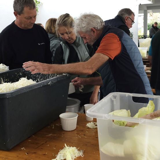 Mehrere Menschen arbeiten gemeinsam an einem Tisch, wobei ein Mann auf einen Behälter mit geriebenem Käse zeigt. Eine Frau und ein Mann stehen daneben und schauen sich den Behälter an.