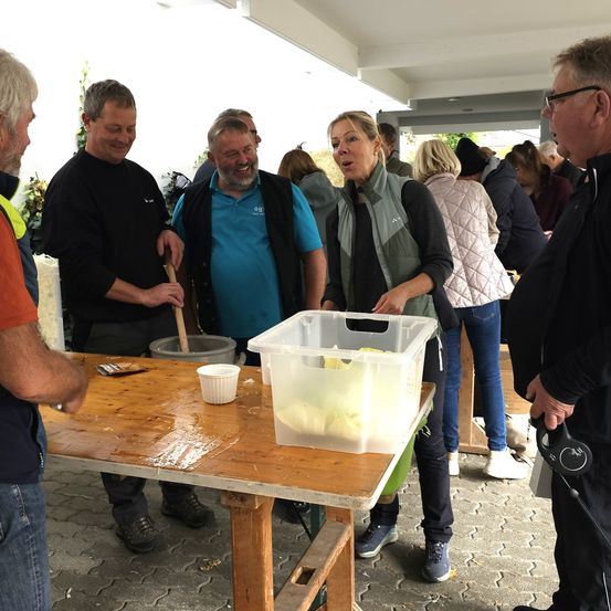 Mehrere Menschen stehen um einen Tisch mit einem Plastikbehälter und lächeln. Eine Frau spricht mit einer anderen Frau mit einem Behälter. Ein Mann lächelt und hält einen Holzstab.