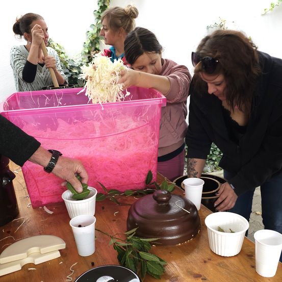 Eine Gruppe von Menschen ist um einen Holztisch versammelt und arbeitet an einem Projekt. Eine Person hält einen pinkfarbenen Plastikbehälter mit zerkleinertem Material. Eine andere Person legt ein Blatt in einen weißen Becher. Auf dem Tisch befinden sich verschiedene Gegenstände, darunter Becher, ein Topf und ein Messer.