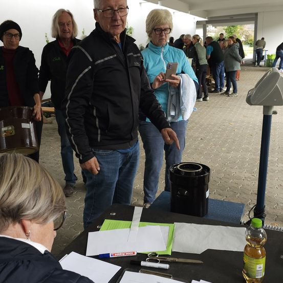 Mehrere Personen versammeln sich in einem Flur, wobei ein Mann auf einen Tisch zeigt. Eine ältere Frau sitzt vor einem Tisch, und ein Fass steht in der Nähe. Papiere, Scheren und eine Flasche befinden sich auf dem Tisch.