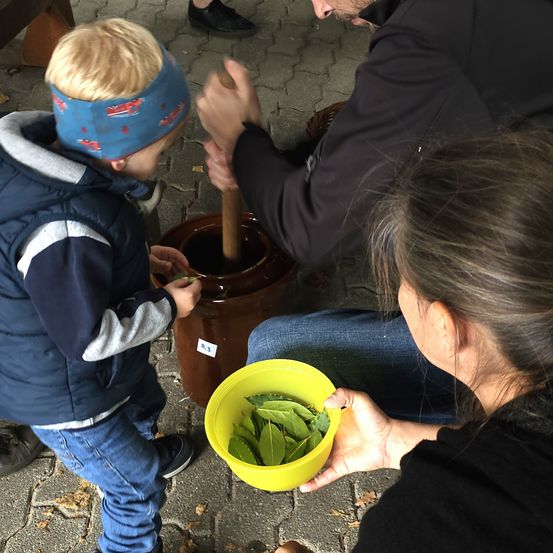 Ein junger Junge fügt Blätter in einen braunen Krug, den eine Frau hält, während ein anderer Erwachsener sie mit einem Holzstab zerkleinert.