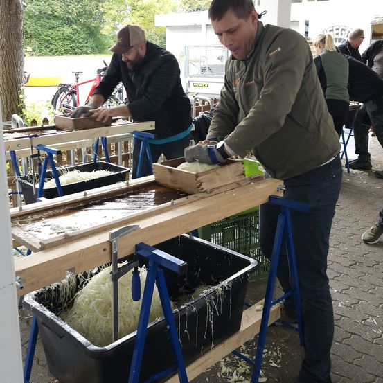 Zwei Männer arbeiten im Freien an einem Holztisch mit einem Wasserbecken. Ein Mann benutzt ein Messer, während der andere eine Kiste hält. Andere Menschen sind im Hintergrund.