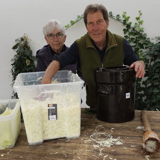 Ein älteres Paar steht lächelnd hinter einem Holztisch. Der Mann hält einen Behälter mit zerkleinertem Kohl. Ein weiterer Behälter steht neben einem schwarzen Topf, und ein hölzerner Schläger liegt auf dem Tisch. Hinter ihnen befindet sich eine mit grünem Laub bedeckte Wand.