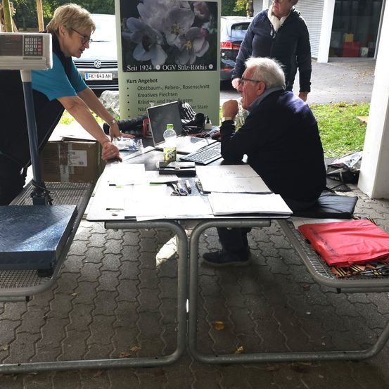Eine Gruppe von Menschen versammelt sich um einen Tisch, wobei eine Person sitzt und einen Laptop benutzt. Eine andere Person steht in der Nähe einer Waage und einer Kiste. Ein Poster mit einer Blume befindet sich hinter ihnen.