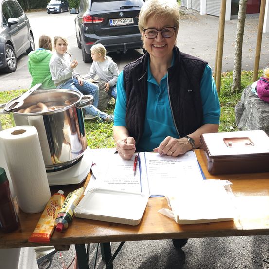 Eine Outdoor-Veranstaltung mit einer Frau in einer Brille, die lächelnd an einem Tisch sitzt. In der Nähe steht ein Topf mit Würstchen, eine Rolle Papiertücher und andere Gegenstände. Hinter ihr sitzen zwei Mädchen auf einem Felsen, und mehrere Autos sind geparkt.
