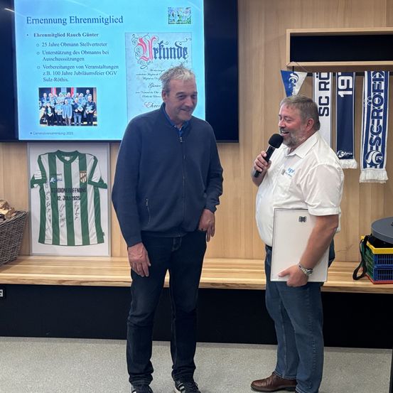 Zwei Männer stehen vor einer Wand, einer hält ein Mikrofon und der andere lächelt. Ein Bildschirm zeigt eine Präsentation über einen Preisträger.