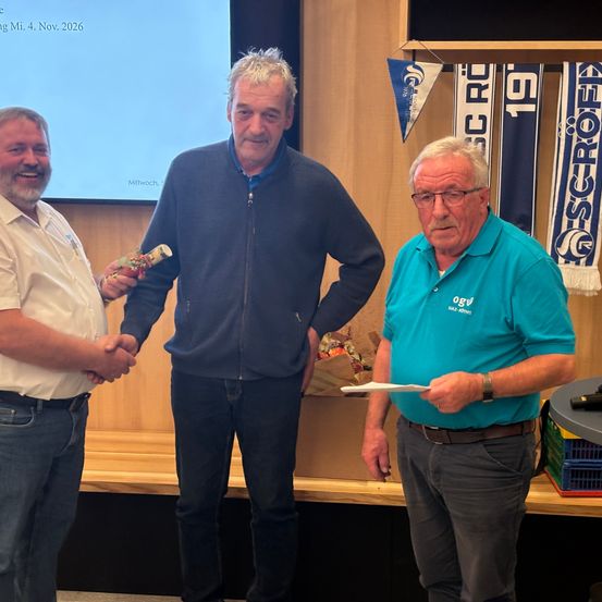 Drei Männer stehen in einem Raum. Einer hält ein verpacktes Geschenk, ein anderer ein Papier und der dritte lächelt. Ein Präsentationsbildschirm ist hinter ihnen sichtbar.