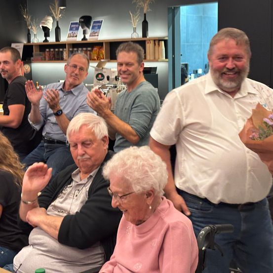 Mehrere Menschen lächeln und klatschen in einem Raum, wobei zwei ältere Personen im Vordergrund sitzen. Ein Mann in einem weißen Hemd hält einen Blumenstrauß.